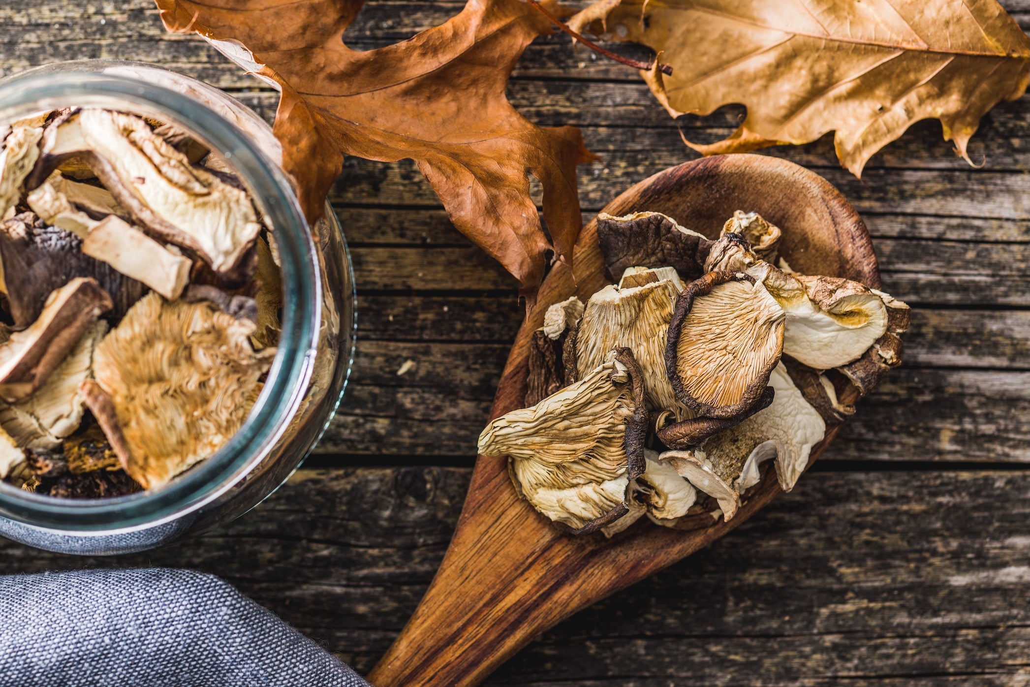 Funghi essiccati disposti su un cucchiaio di legno, con barattolo e foglie autunnali su fondo rustico
