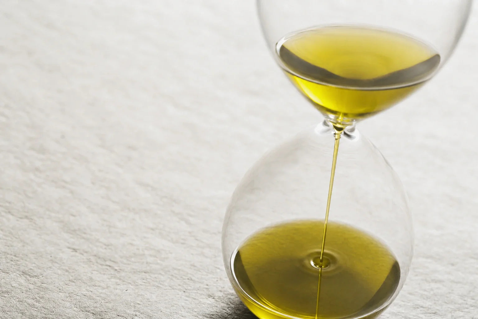 Clessidra di vetro con olio dorato che scorre lentamente da un’ampolla all’altra, su fondo chiaro.