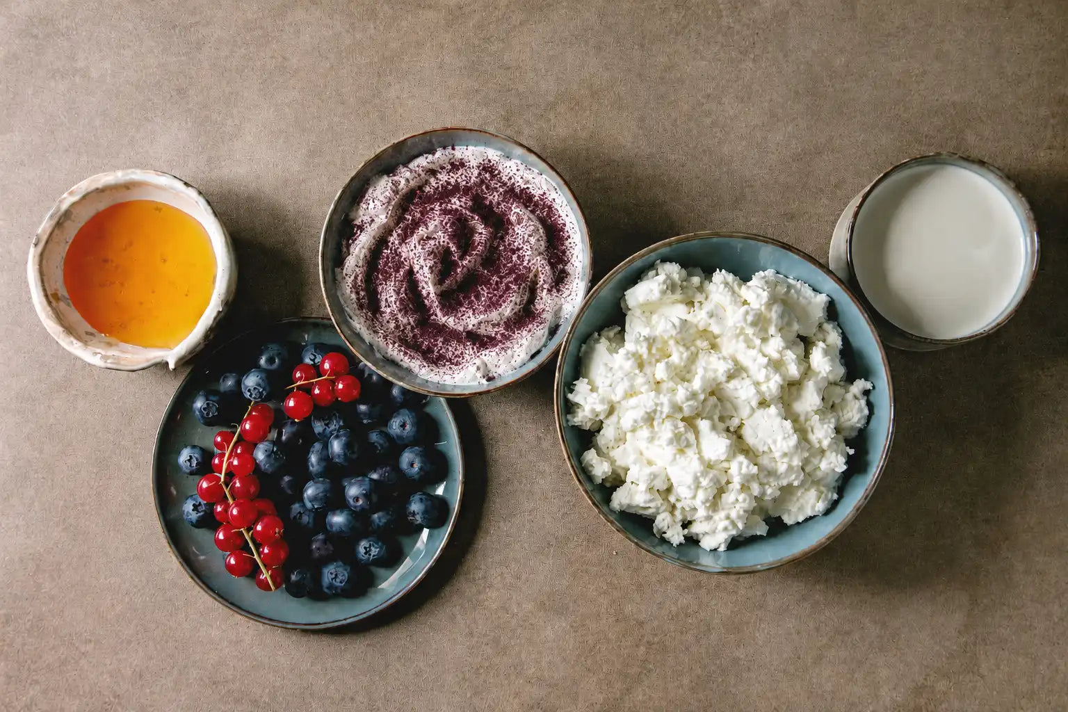 Ciotola di yogurt bianco spolverato con polvere di vinaccia, accompagnata da frutti di bosco, miele, ricotta e latte su fondo neutro.