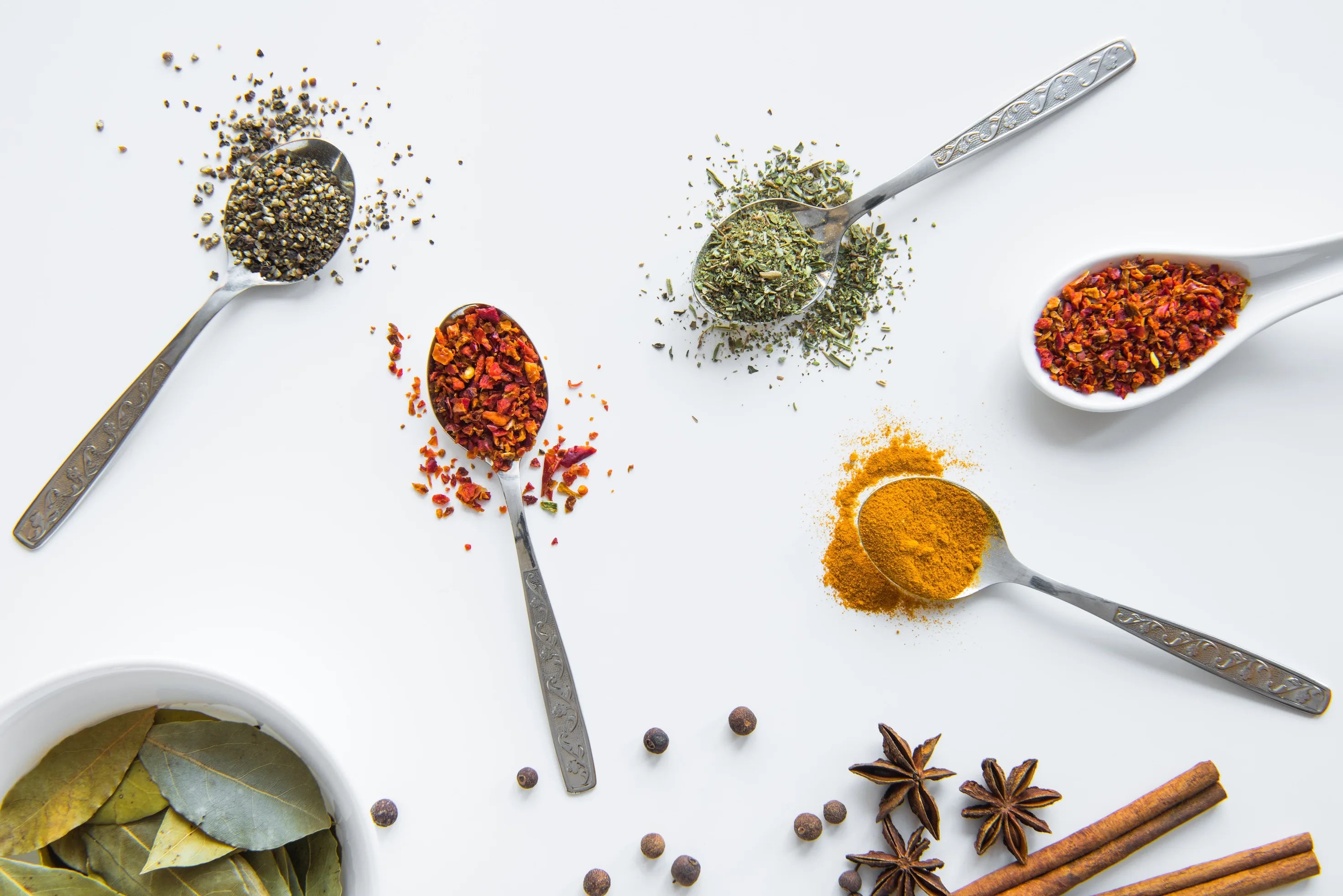 Assortimento di spezie ed erbe aromatiche in cucchiai su fondo bianco, con peperoncino, curcuma, pepe, alloro, anice stellato e cannella.